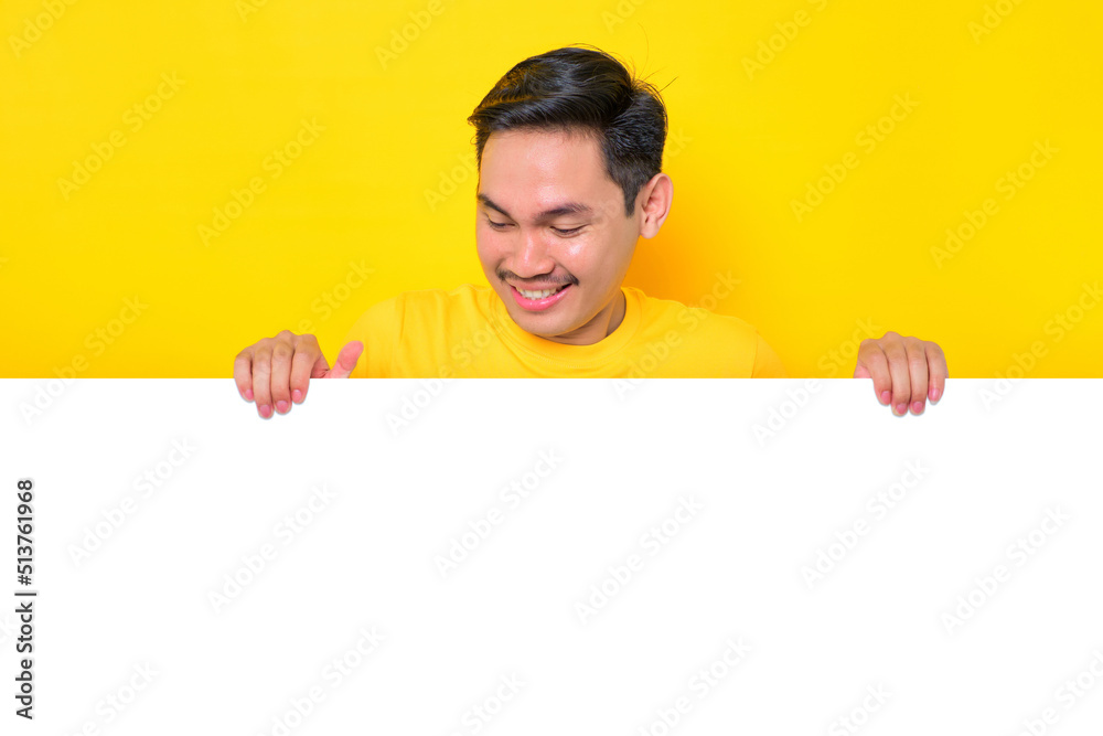 Cheerful young Asian man in casual t-shirt looking down at white advertisement board isolated on yellow background. Promotion billboard concept