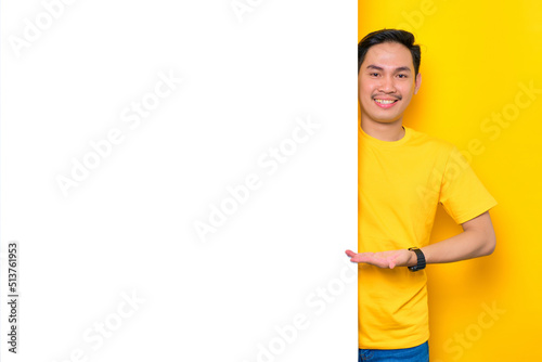 Cheerful young Asian man in casual t-shirt presenting something on white advertisement board isolated on yellow background. Promotion billboard concept
