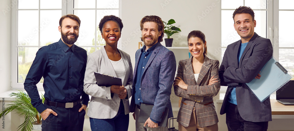 Obraz premium Happy diverse business team standing in the office. Five cheerful confident successful people in smart casual suits smiling and posing for a group photo all together. Banner. Work and success concept