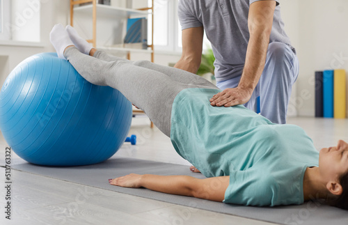 Young woman lying on rubber mat and doing special exercises with fit ball. Specialist at modern physiotherapy and rehabilitation center helping female patient do physical exercises with stability ball photo