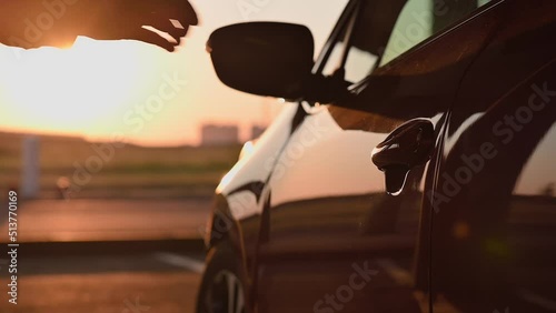 A young attractive girl in light clothes opens the door of red car and gets behind the wheel at sunset. Cinematic slow motion footage with rays of the setting sun. Girl driver, lifestyle, travel