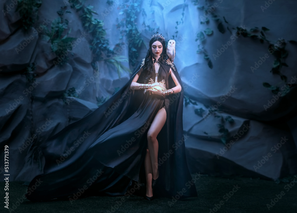 Fototapeta premium Fairy woman elf queen in black fantasy sexy long dress fly in wind cape flutter waving flowing around witch Princess goth girl sharp ears golden crown. old style cave. Spell light in hand dark magic 