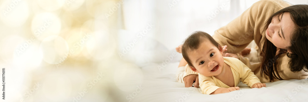 baby girl with asian mom smiling baby happily playing together in bed in the bedroom at home web banner with copy space on left baby girl with asian mom smiling baby happily playing together in bed in the bedroom at home web banner with copy space on left