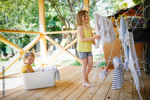 Cute European sibling children hang laundry together on veranda in backyard in summer. Helping children with household routines, relationships and sibling games in family photo