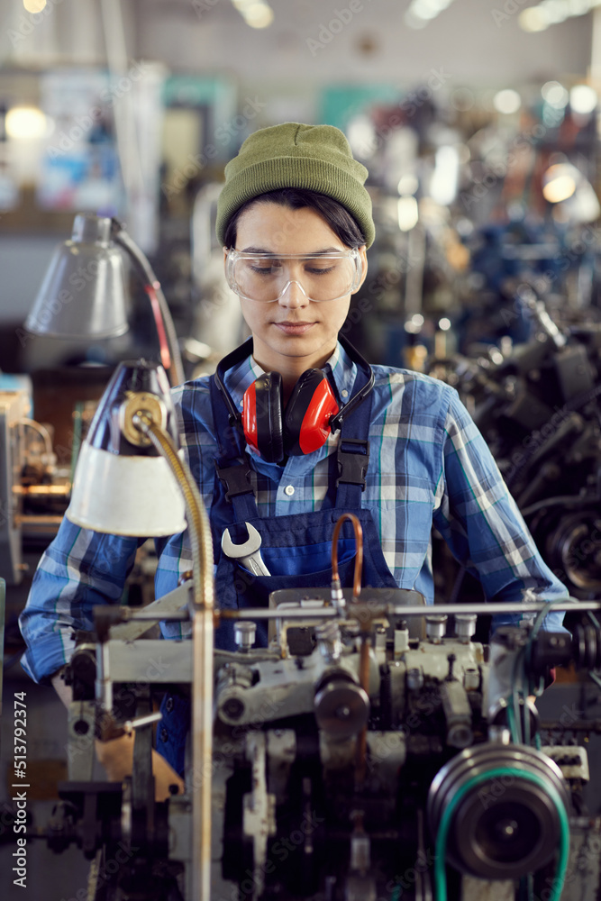 Serious concentrated young female mechanic in protective goggles and hipster hat standing in industrial workshop and repairing machine