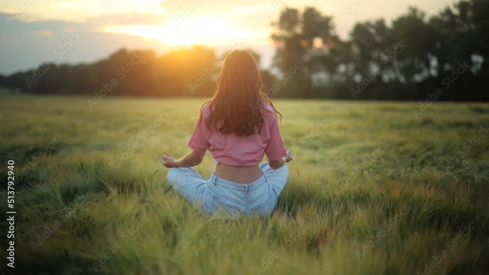 Back view of slender young woman sitting on yoga mat in lotus position in the field. Girl holds a meditation session against the backdrop of an incredible landscape Back view of slender young woman sitting on yoga mat in lotus position in the field. Girl holds a meditation session against the backdrop of an incredible landscape