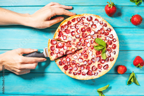Woman's hands cut the homemade strawberry custard tart decorated with strawberry on blue wooden background. Top view
