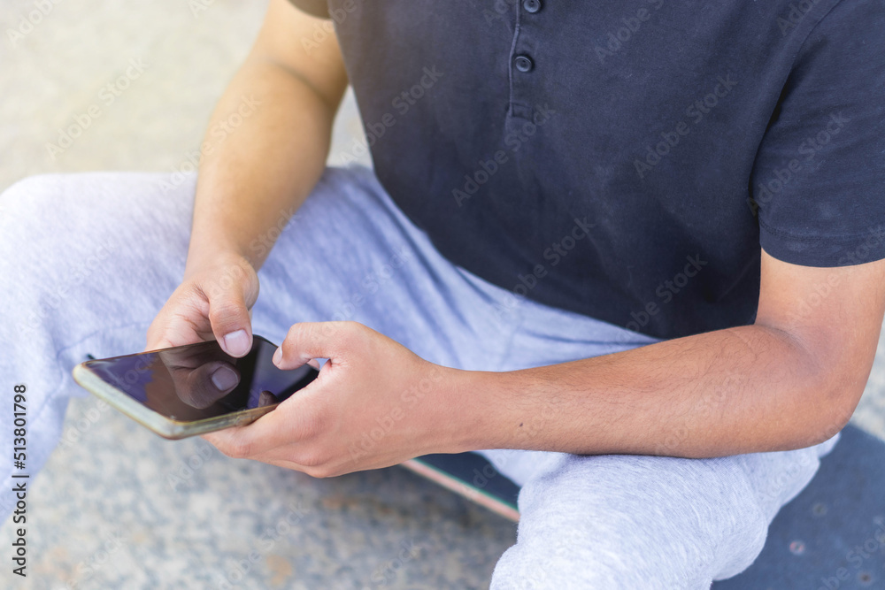 Close photo of dark-skinned guy sitting on a skateboard and unlocking his cell phone.