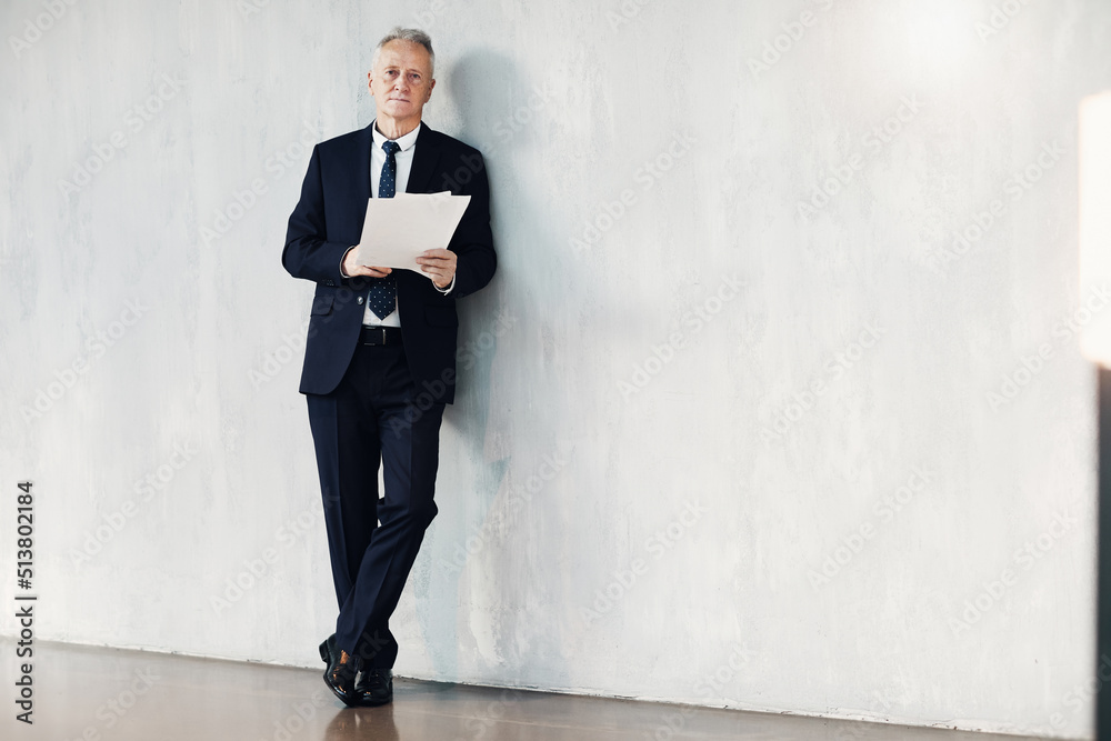 Portrait of serious successful senior businessman in black suit leaning on wall in lobby and reading papers Portrait of serious successful senior businessman in black suit leaning on wall in lobby and reading papers