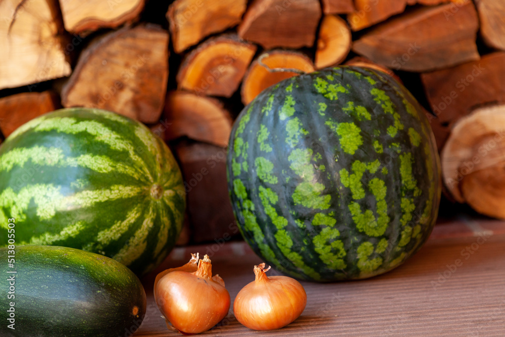 Watermelons and vegetables lie on floor in front of firewood..