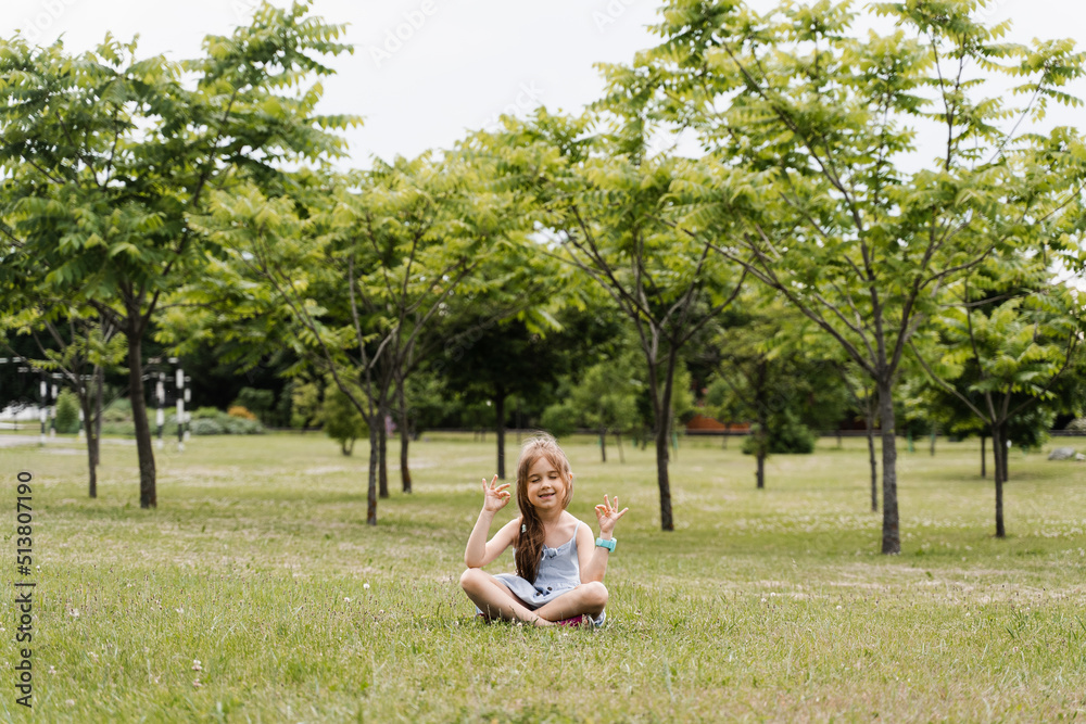 Adorable child girl is sitting and meditate in lotus pose in the park. Relaxation and meditation of kids outdoor. Lifestyle of calm toddler. Enjoy life. Adorable child girl is sitting and meditate in lotus pose in the park. Relaxation and meditation of kids outdoor. Lifestyle of calm toddler. Enjoy life.