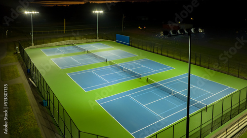 Evening aerial photo of outdoor blue tennis courts with pickleball lines with lights turned on. 