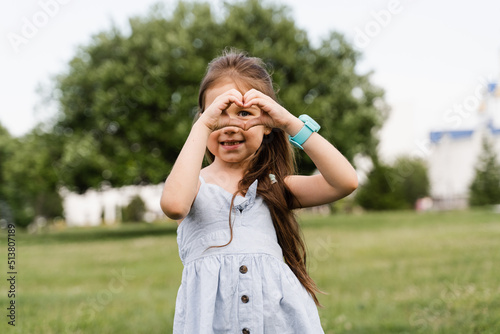 Adorable child girl showing heart with her hands. Cute toddler shows peace and love. Happy kid smiling and posing in green park.