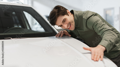 Cheerful young Caucasian man touching his brand new car, buying vehicle at dealership store, banner with free space