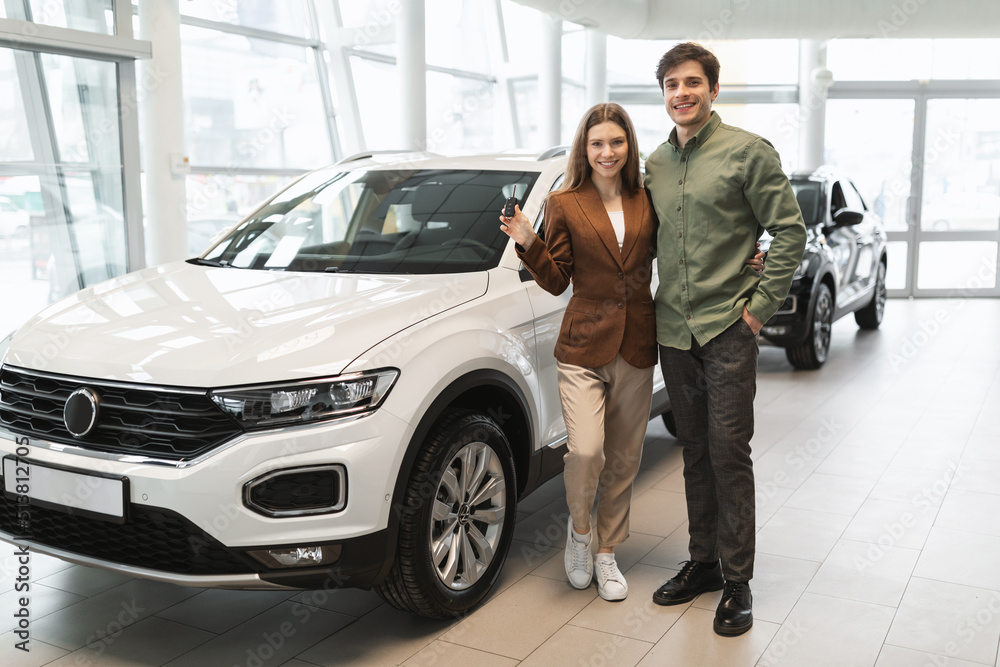 Full length of cheery young couple showing key to their new car at automobile dealership store, free space