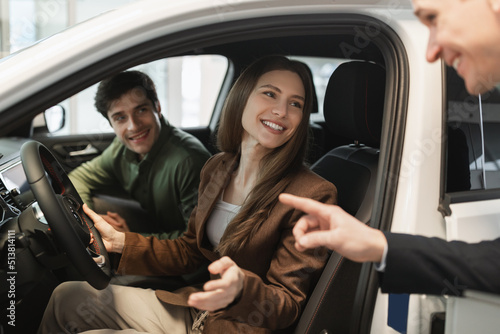 Positive millennial couple test driving new car, salesman helping them make choice at auto dealership
