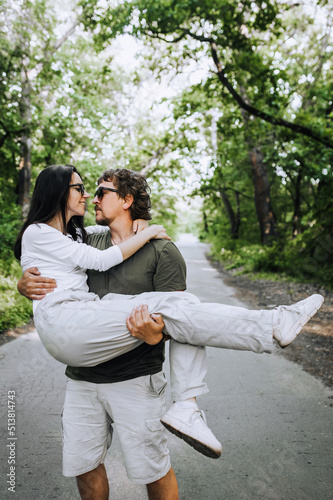 A bearded long-haired stylish man holds his beloved hippie brunette woman in sunglasses in his arms and circles her in nature in the park. Portrait, photo of happy and smiling newlyweds.