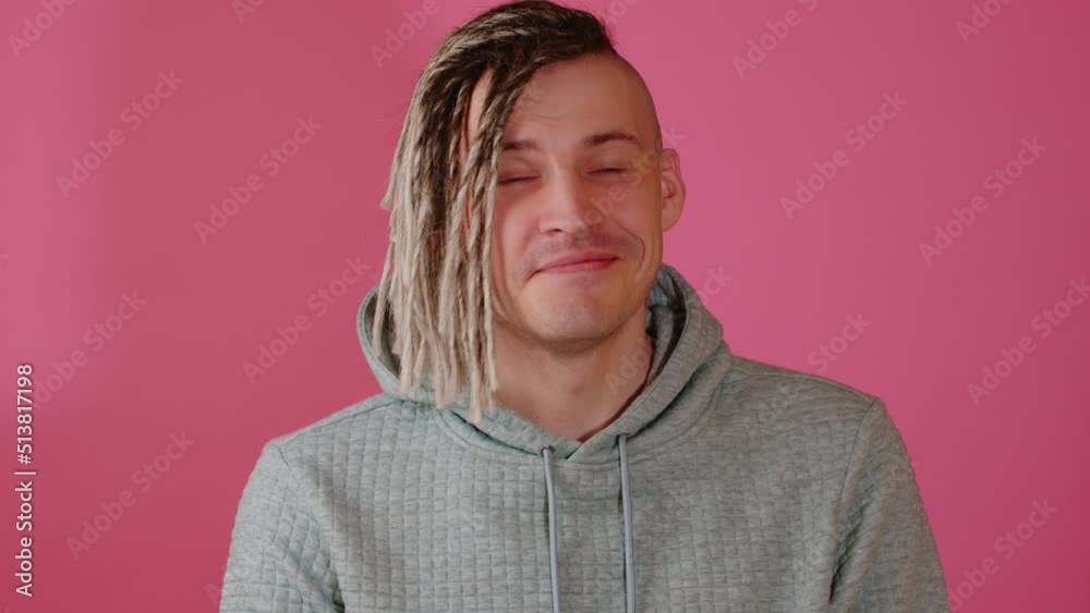 Young handsome man with blonde dreadlocks looking at camera, smiling on pink background. Attractive happy guy in grey hoodie with positive emotions posing in studio. Young handsome man with blonde dreadlocks looking at camera, smiling on pink background. Attractive happy guy in grey hoodie with positive emotions posing in studio.