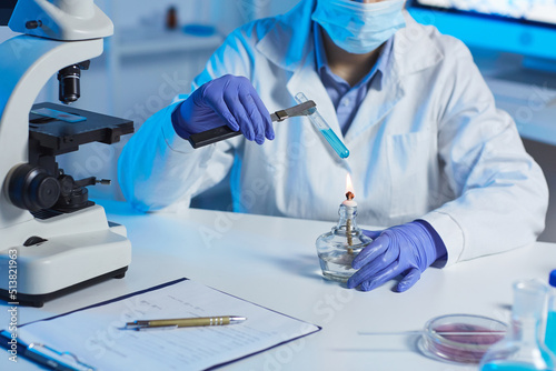 Close-up of unrecognizable chemist in rubber gloves sitting at table and heating test tube filled with reagents over gas burner