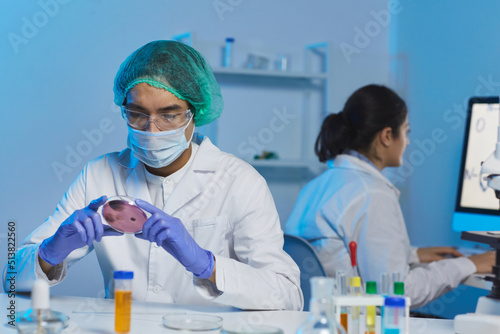 Serious concentrated young Asian male bacteriologist in surgical cap and mask sitting at desk and analyzing bacteria cell in petri dish photo