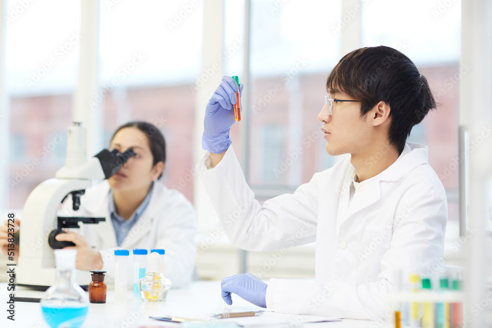 Serious young Asian lab employee in white coat sitting at table with laboratory tools and looking at liquid in test tube Serious young Asian lab employee in white coat sitting at table with laboratory tools and looking at liquid in test tube
