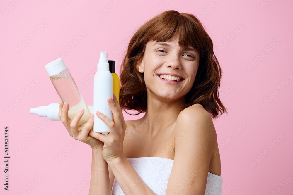 a happy young woman stands on a pink background, wrapped in a white towel and holding a set of jars with facial care cosmetics, smiling broadly and squinting her eyes a happy young woman stands on a pink background, wrapped in a white towel and holding a set of jars with facial care cosmetics, smiling broadly and squinting her eyes