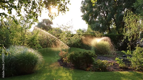 Landscape automatic garden watering system with different sprinklers installed under turf. Landscape design with lawn hills and fruit garden irrigated with smart autonomous sprayers at sunset time