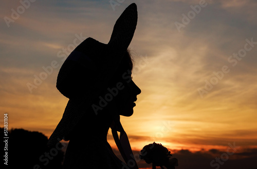 Close up silhouette of a woman in a hat at sunset. Image against the sun and orange sky. High quality horizontal photo
