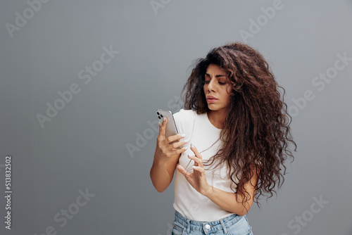 Confused curly Latin woman in white shirt using phone, sms from fakers, reading bad stress gossip news, posing isolated on gray background, studio portrait. People emotion concept. Mock up copy space photo