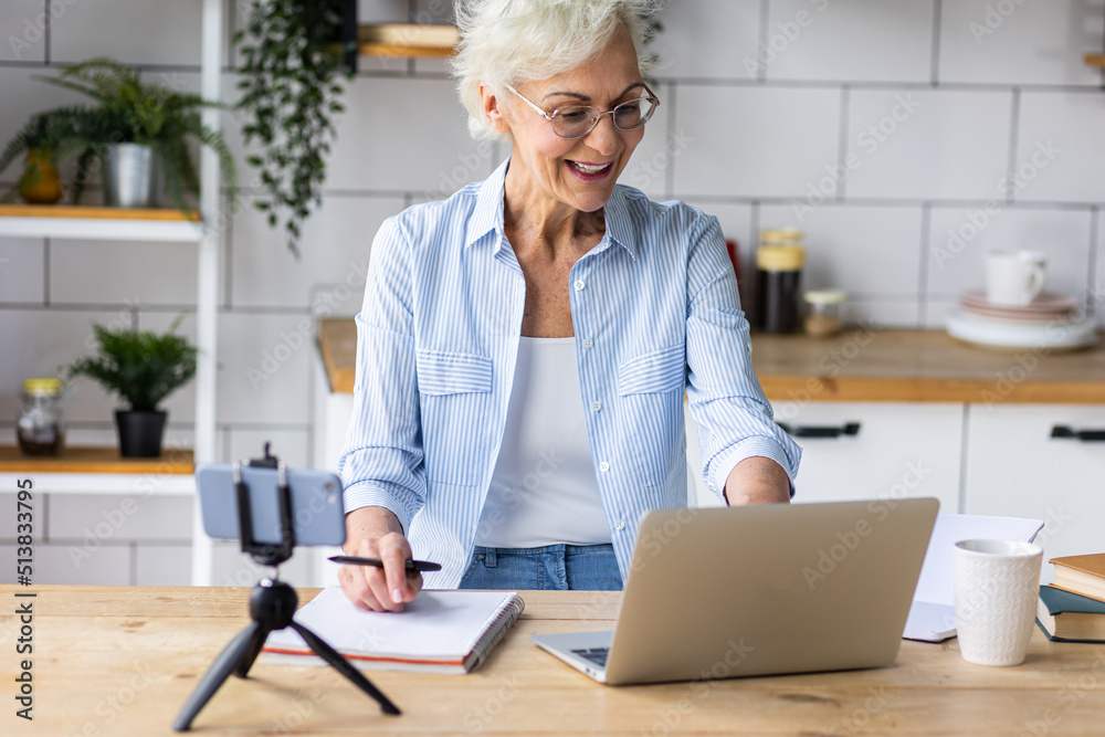 Attractive senior woman working at home in the kitchen using her laptop for conference call. Concept of mature female using internet, modern technology for learning, shopping, freelance job Attractive senior woman working at home in the kitchen using her laptop for conference call. Concept of mature female using internet, modern technology for learning, shopping, freelance job