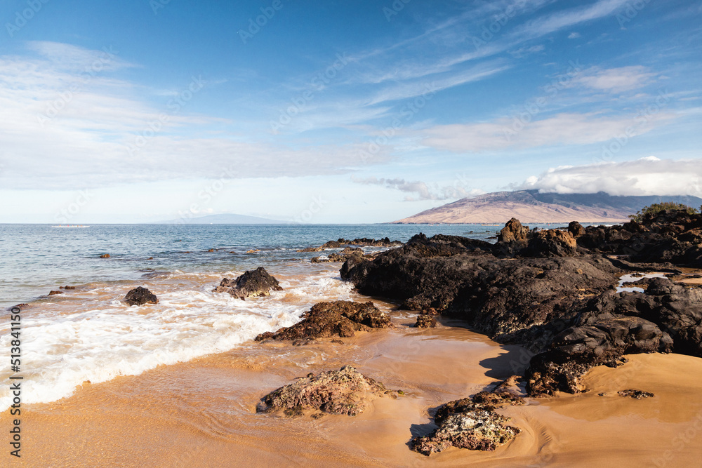 A rocky beach in Maui Hawaii with sand, waves and a mountain in the distance