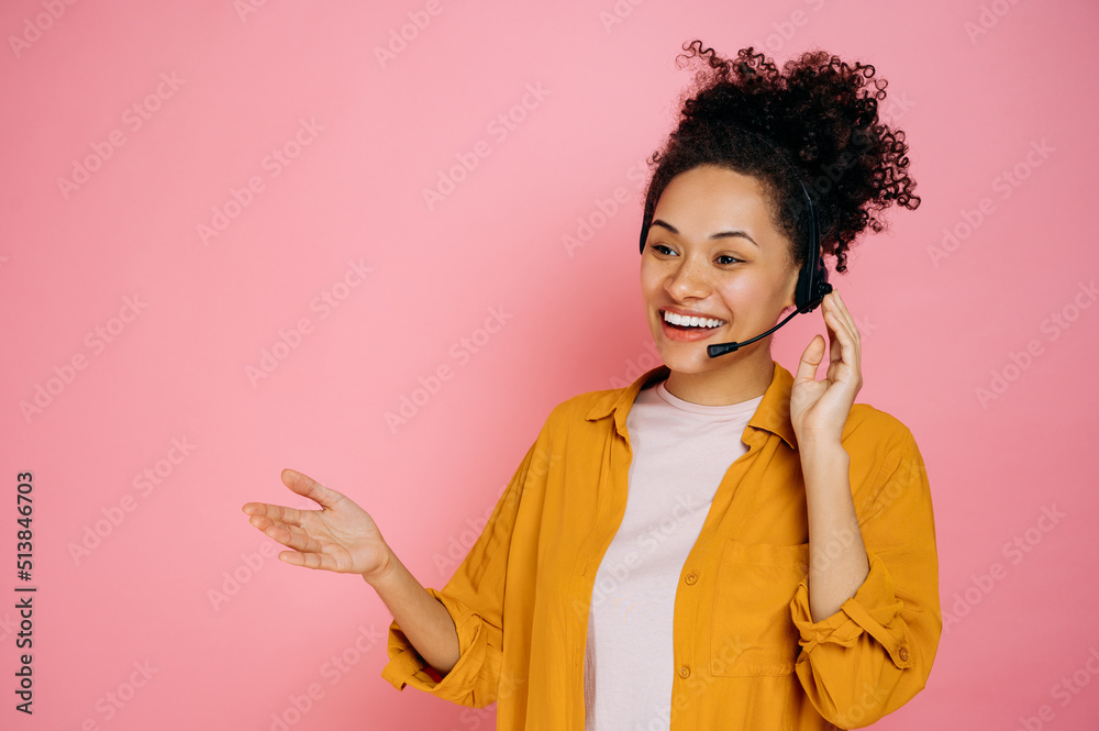 Positive african american curly haired young woman with headset, customer support service, call center operator, expert, advises clients, standing on isolated pink background, smiling friendly - obrazy, fototapety, plakaty 
