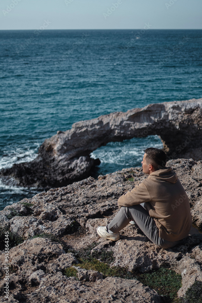 Man from behind is sitting at the edge and looking at sea cave arch bridge Man from behind is sitting at the edge and looking at sea cave arch bridge