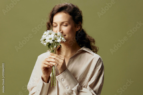 Smiling cute curly beautiful female in linen casual shirt hold chamomiles flowers near face enjoying smell posing isolated on over olive green pastel background. Nature is beauty concept. Copy space