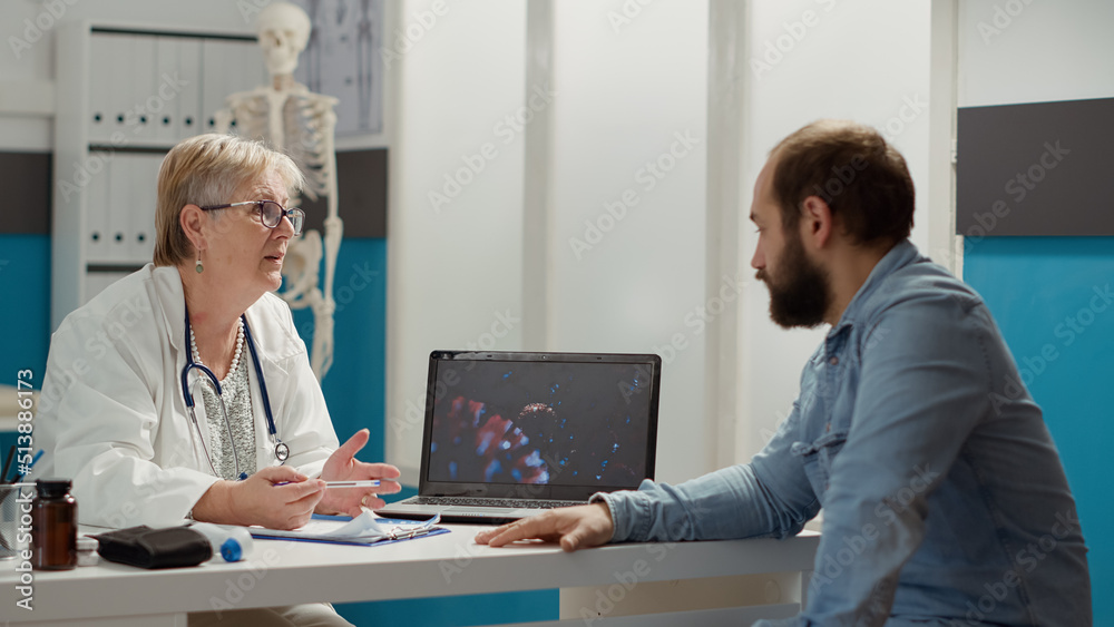 Female medic looking at coronavirus illustration on laptop with sick patient, talking about medication and prevention. Virus animation on display to cure disease in medical cabinet. Female medic looking at coronavirus illustration on laptop with sick patient, talking about medication and prevention. Virus animation on display to cure disease in medical cabinet.