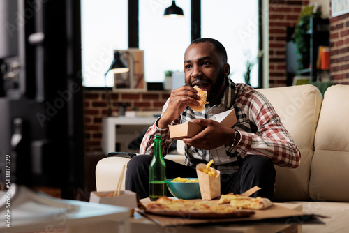 African american man eating burger from takeaway delivery and watching movie on television program. Enjoying fast food takeout in package with bottle of beer in front of tv film, having fun.