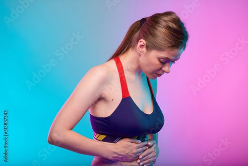 Woman hand on stomach, abdominal pain and digestive disorder. Girl looking down. Female fitness portrait isolated on neon multicolor background.