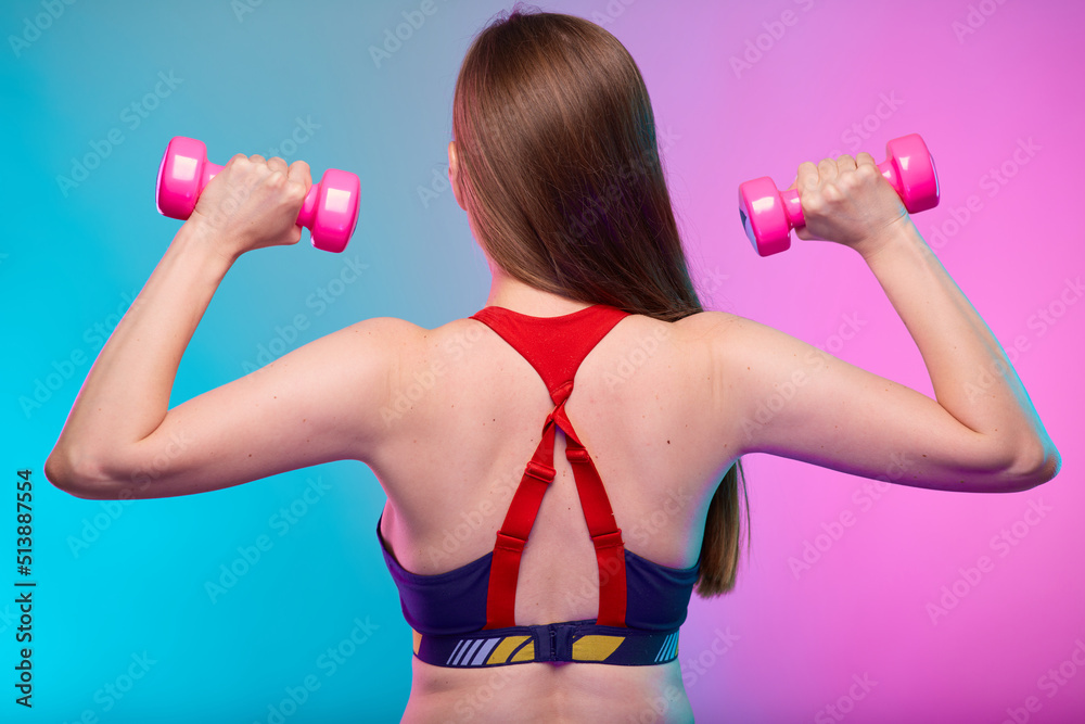 Sporty woman with long hair in fitness sportswear holding pink dumbbells back view. Female fitness portrait isolated on neon multicolor background. Sporty woman with long hair in fitness sportswear holding pink dumbbells back view. Female fitness portrait isolated on neon multicolor background.