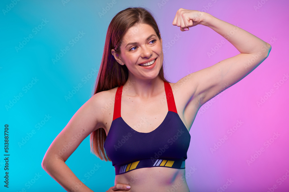 Fototapeta premium Happy young sporty woman in fitness sportswear flexes arm and demonstrates muscles, looking away. Female fitness portrait isolated on neon multicolor background.