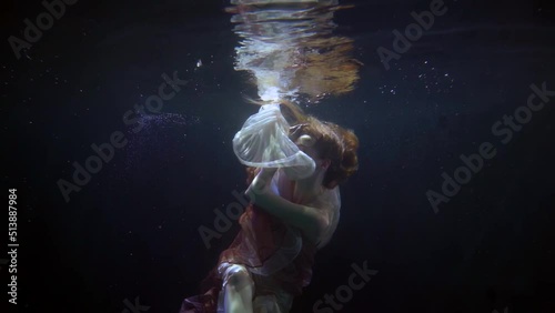 a red-haired woman hovers in the dark water. she is wrapped in white and red translucent fabrics