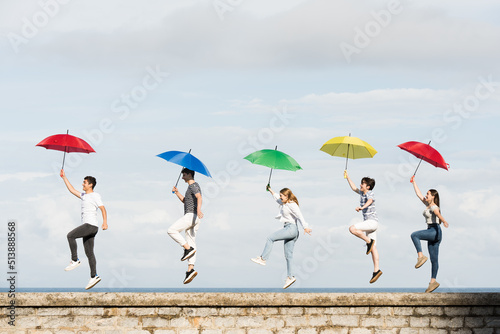 Side view of teenager friends jumping and dancing together on a wall while holding colorful umbrellas