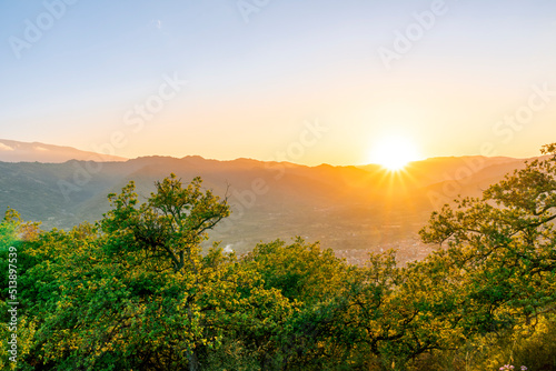 amazing highland landscape with scenic view from mountain with green branches and leaves on sides to a valley town with majectic mountains and scenic cloudy sunset on background