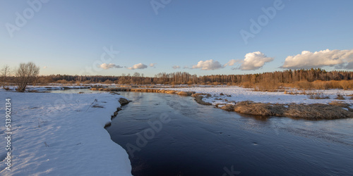 March sunny evening by the river. Blue sky over the horizon. A picturesque landscape, early spring, a river with snow-covered banks, dry grass and bushes. The first thaws, the snow is melting