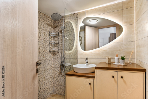 Interior design of hotel or apartment or home standard modern bathroom with silver rain head, hand held shower, sink, mirror with backlight, beige colors. 