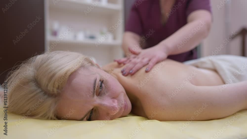 Smiling relaxed woman lying in beauty spa with unrecognizable blurred masseur rubbing back in slow motion. Portrait of confident Caucasian client enjoying massage looking away Smiling relaxed woman lying in beauty spa with unrecognizable blurred masseur rubbing back in slow motion. Portrait of confident Caucasian client enjoying massage looking away