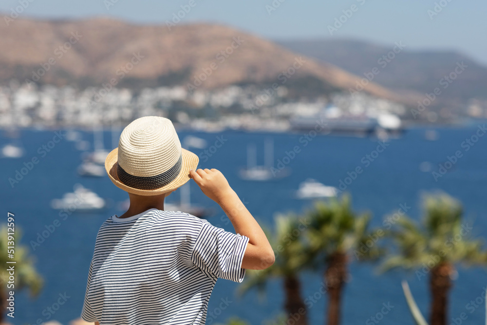 Back view of boy wearing striped t-shirt and summer hat watching the sea from above.