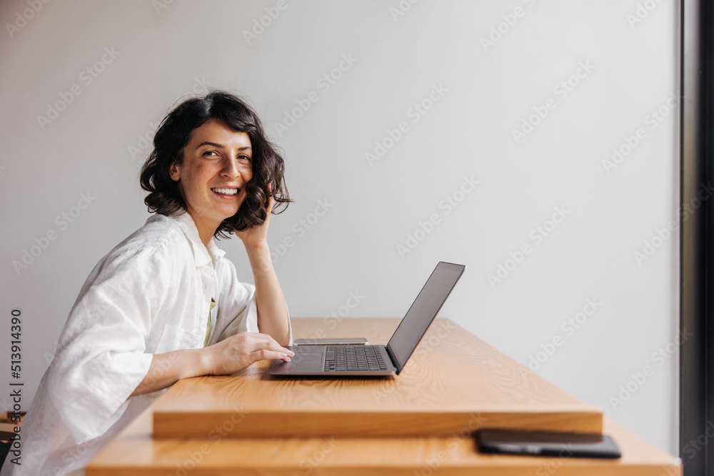 Happy young caucasian brunette girl looks at camera sitting at laptop in room. Woman wearing shirt checks her mailbox. Concept of use Happy young caucasian brunette girl looks at camera sitting at laptop in room. Woman wearing shirt checks her mailbox. Concept of use