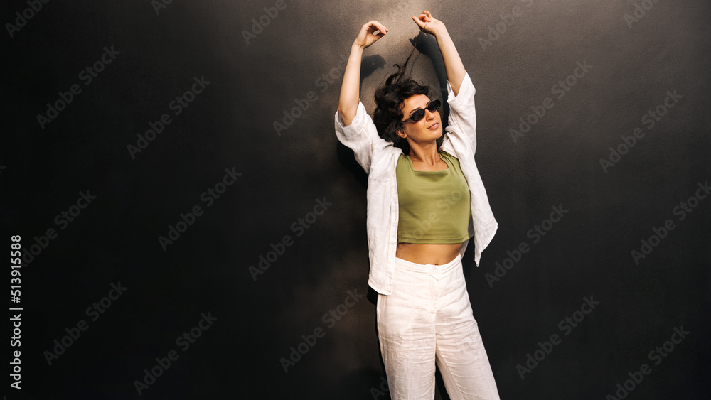 Relaxed caucasian young woman stretching arms above her head standing on black background with space for text. Brunette wears sunglasses and casual clothes. Rest time concept Relaxed caucasian young woman stretching arms above her head standing on black background with space for text. Brunette wears sunglasses and casual clothes. Rest time concept