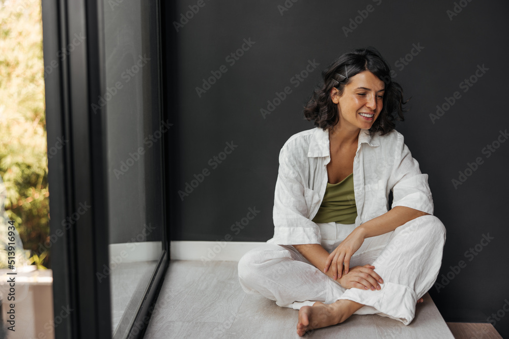 Positive young caucasian woman in good mood sits on floor near window in morning. Brunette with wavy hair wears shirt and pants. People emotions, lifestyle and fashion concept.
