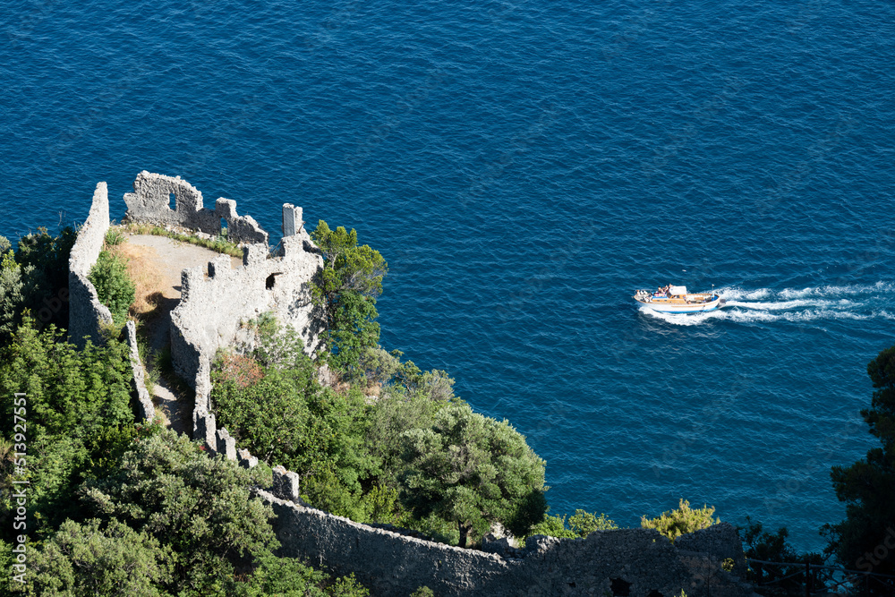 View from above, stunning view of a gozzo boat with tourists on board sailing on a blue water. Defocused Torre dello Ziro in the foreground. Amalfi Coast, Italy.